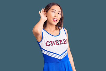 Young beautiful chinese girl wearing cheerleader uniform smiling with hand over ear listening an...