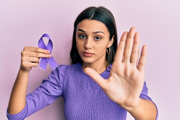 Young hispanic woman holding purple ribbon awareness with open hand doing stop sign with serious...