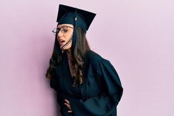 Young hispanic woman wearing graduation cap and ceremony robe with hand on stomach because nausea,...