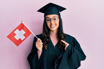Young hispanic woman wearing graduation uniform holding switzerland flag smiling happy pointing with hand and finger