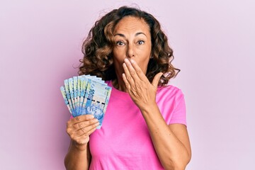 Middle age hispanic woman holding south african 20 rand banknotes covering mouth with hand, shocked and afraid for mistake. surprised expression