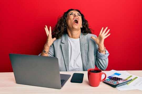 Beautiful middle age woman working at the office drinking a cup of coffee crazy and mad shouting and yelling with aggressive expression and arms raised. frustration concept.
