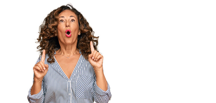 Middle age hispanic woman wearing casual clothes amazed and surprised looking up and pointing with fingers and raised arms.