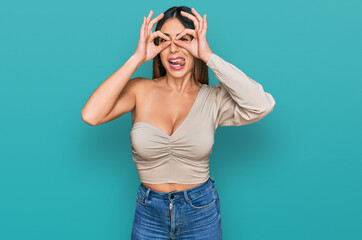 Young hispanic woman wearing casual clothes doing ok gesture like binoculars sticking tongue out, eyes looking through fingers. crazy expression.