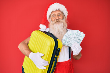 Old senior man with grey hair and long beard wearing santa claus costume holding dollars and suitcase smiling with a happy and cool smile on face. showing teeth.