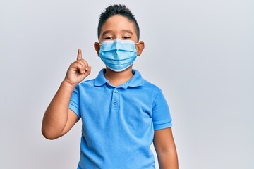Little boy hispanic kid wearing medical mask smiling with an idea or question pointing finger up with happy face, number one