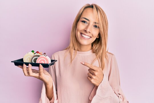 Beautiful blonde woman holding cake sweets smiling happy pointing with hand and finger
