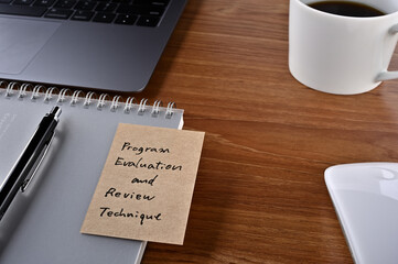 On the desk there is a laptop, a cup of coffee, and a sticky note with the word financial term Program Evaluation and Review Technique.