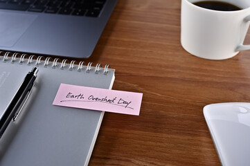 There's a laptop, a cup of coffee and a notebook with a sticky note stuck to it that says Earth Overshoot Day.