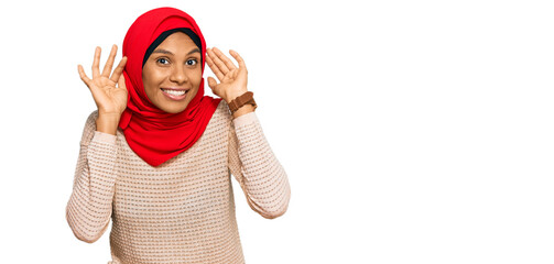 Young african american woman wearing traditional islamic hijab scarf smiling pulling ears with...