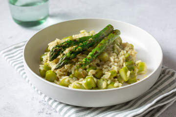 Tasty risotto with grilled asparagus on white plate, grey background, vertical with green glass, horizontal