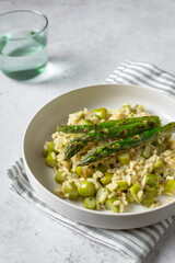 Tasty risotto with grilled asparagus on white plate, grey background, vertical with green glass
