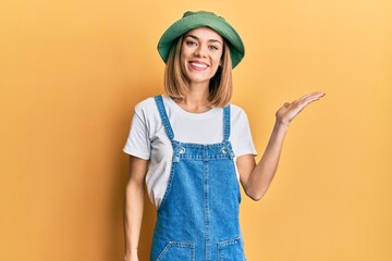 Young caucasian blonde woman wearing denim jumpsuit and hat with 90s style smiling cheerful presenting and pointing with palm of hand looking at the camera.