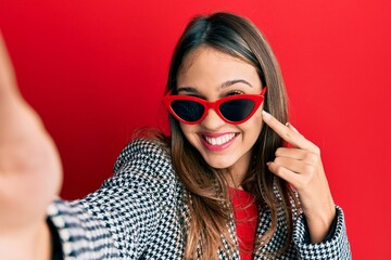 Young brunette woman taking a selfie photo wearing sunglasses smiling happy pointing with hand and finger