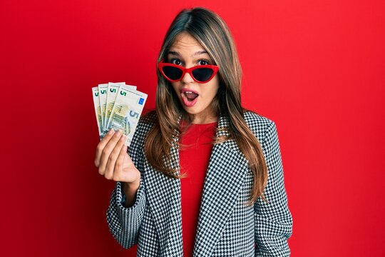 Young brunette woman holding bunch of 5 euro banknotes scared and amazed with open mouth for surprise, disbelief face