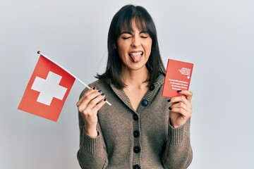 Young hispanic woman holding swiss flag and passport sticking tongue out happy with funny...