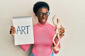 Young african american girl holding wooden manikin and art word on notebook winking looking at the...