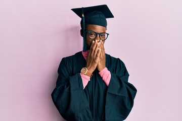 Young african american girl wearing graduation cap and ceremony robe laughing and embarrassed giggle covering mouth with hands, gossip and scandal concept