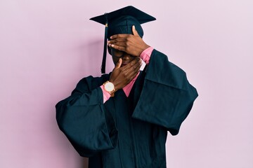 Young african american girl wearing graduation cap and ceremony robe covering eyes and mouth with hands, surprised and shocked. hiding emotion