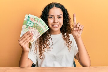 Teenager hispanic girl holding 50 hong kong dollars banknotes smiling with an idea or question pointing finger with happy face, number one