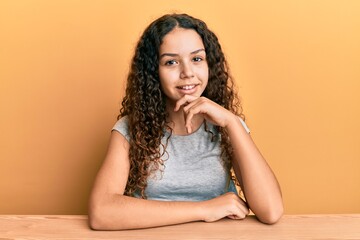 Teenager hispanic girl wearing casual clothes sitting on the table looking confident at the camera smiling with crossed arms and hand raised on chin. thinking positive.