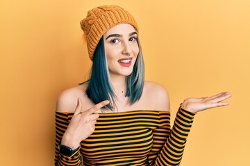 Young modern girl wearing wool hat amazed and smiling to the camera while presenting with hand and pointing with finger.