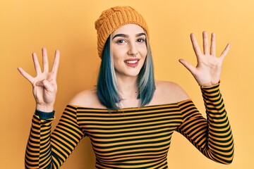 Young modern girl wearing wool hat showing and pointing up with fingers number nine while smiling...