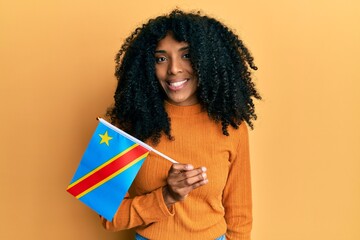 African american woman with afro hair holding democratic republic of the congo flag looking positive and happy standing and smiling with a confident smile showing teeth