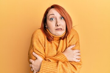 Beautiful redhead woman wearing casual winter sweater over yellow background shaking and freezing for winter cold with sad and shock expression on face