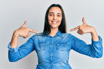 Young hispanic woman wearing casual denim jacket looking confident with smile on face, pointing...