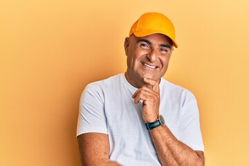 Mature middle east man with mustache wearing casual white tshirt and yellow cap smiling looking confident at the camera with crossed arms and hand on chin. thinking positive.