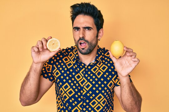 Handsome hispanic man with beard holding lemon in shock face, looking skeptical and sarcastic, surprised with open mouth