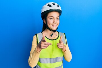 Beautiful brunette little girl wearing bike helmet and reflective vest pointing fingers to camera with happy and funny face. good energy and vibes.