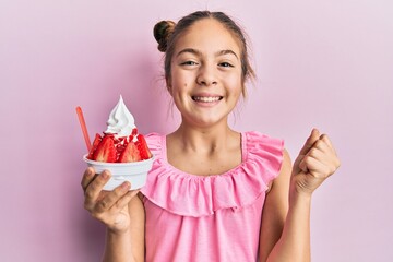 Beautiful brunette little girl eating strawberry ice cream screaming proud, celebrating victory and...