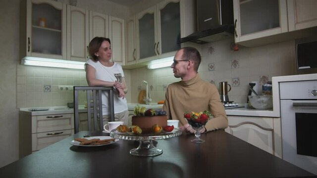 Adult happy couple talks at kitchen. Cake and cookies on table