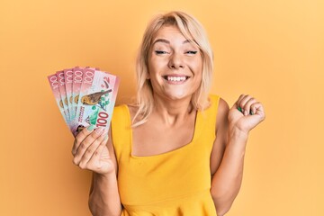 Young blonde girl holding 100 new zealand dollars banknote screaming proud, celebrating victory and success very excited with raised arm