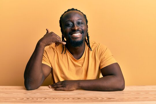Handsome young black man wearing casual clothes sitting on the table smiling doing phone gesture with hand and fingers like talking on the telephone. communicating concepts.