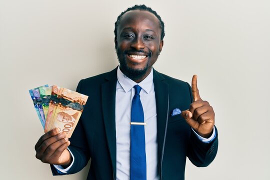 Handsome young black man wearing business suit and tie holding canadian dollars smiling with an idea or question pointing finger with happy face, number one