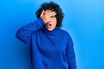 Fototapeta na wymiar Young hispanic woman with curly hair wearing casual winter sweater peeking in shock covering face and eyes with hand, looking through fingers afraid