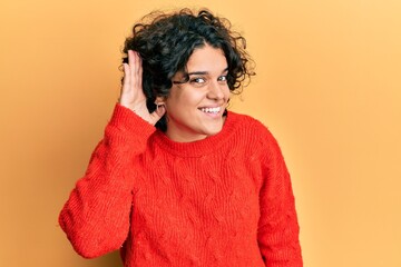 Young hispanic woman with curly hair wearing casual winter sweater smiling with hand over ear listening an hearing to rumor or gossip. deafness concept.