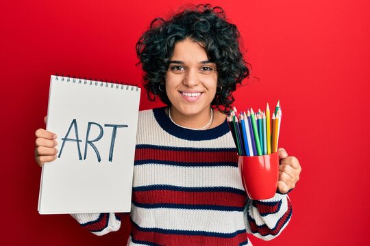 Young hispanic woman with curly hair holding canvas book and colored pencils smiling with a happy and cool smile on face. showing teeth.