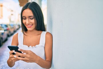 Young latin girl smiling happy using smartphone leaning on the wall.