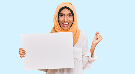 Young brunette arab woman wearing traditional islamic hijab holding banner screaming proud, celebrating victory and success very excited with raised arms