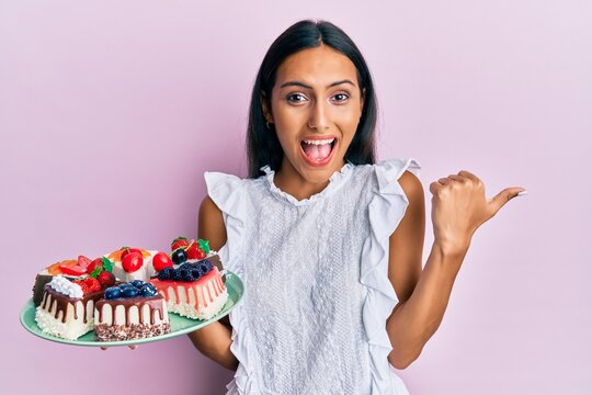 Young brunette woman holding cake slices pointing thumb up to the side smiling happy with open mouth