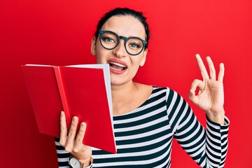 Young caucasian woman wearing glasses covering mouth with book doing ok sign with fingers, smiling friendly gesturing excellent symbol