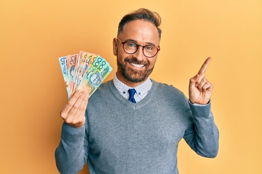 Handsome middle age man holding australian dollars smiling happy pointing with hand and finger to the side