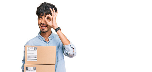 Young hispanic man holding delivery package smiling happy doing ok sign with hand on eye looking through fingers