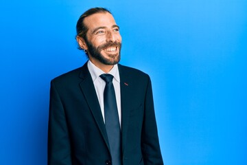 Attractive man with long hair and beard wearing business suit and tie looking away to side with smile on face, natural expression. laughing confident.