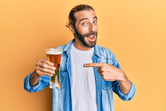 Attractive man with long hair and beard drinking a pint of beer smiling happy pointing with hand and finger