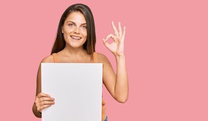 Young caucasian woman holding blank empty banner doing ok sign with fingers, smiling friendly gesturing excellent symbol